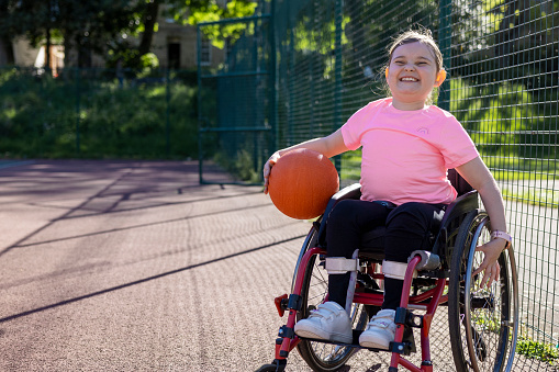 A portrait of a young girl who is a wheelchair user, holding a basketball on a sports court in a public park in Newcastle upon Tyne, England. She is looking at the camera with a big smile on her face.