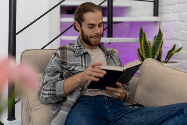 istock Man reading book while sitting on cough at home, enjoying interesting novel. 1488891803