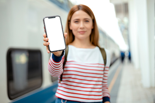 App ad. Woman passenger holding smartphone with white blank device screen in hand close up to camera in a train station. Gadget with empty free space for mockup, banner isolated, selective focus