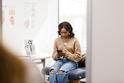The female university student reads a text message on her phone while waiting for the doctor.