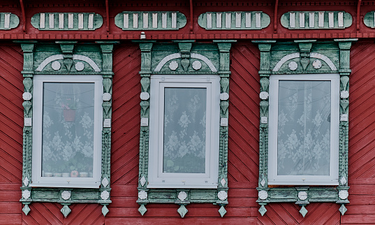 Three windows with carved wooden green architraves on red painted, sheathed with wooden slats facade of typical rural house. Nizhny Novgorod region, Russia. Architecture concept.