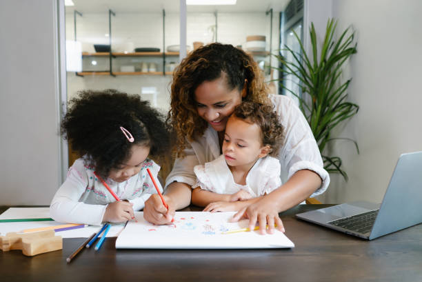 Smiling African American family mother with children sit at kitchen table together paint. Single mother helps the child girl draw. Young Black mother do freelance the desk with little child at home.
