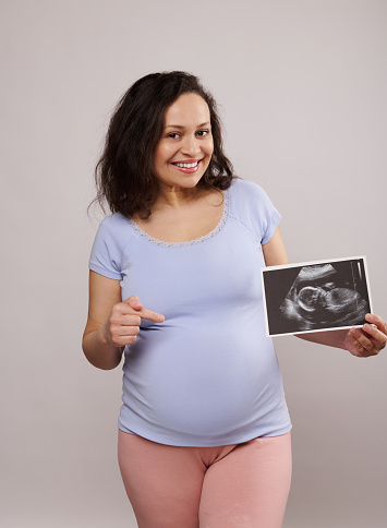 Happy adult pregnant woman, smiling looking at camera, showing her newborn baby sonography and pointing at her belly over isolated isolated white background. Pregnancy. Ultrasound scan image of a baby