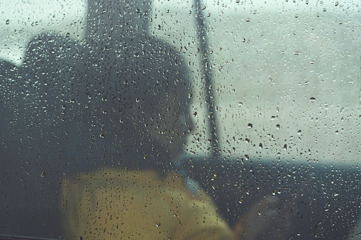 Sad young woman sitting lonely and depressed inside a car through raindrops on window