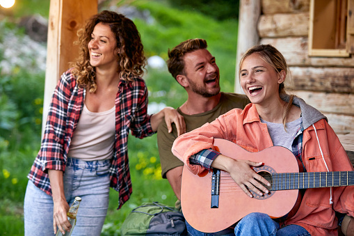 Three cheerful young friends enjoying vacation day and playing guitar in front of wooden cabin. Beuatiful moments. Vacation, holiday, togetherness concept.