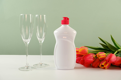 A white plastic bottle of waching dishes cleaning agent with space for text and red flowers, two glasses on a white green background.