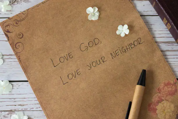 Love God, love your neighbor handwritten text verse on vintage paper with closed bible book and pen on wooden table. Christian faith and obedience to Jesus Christ, the greatest commandment concept.