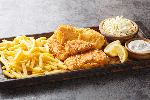 Close up battered fish with coleslaw, French fries, tartar sauce and lemon on the wooden board on the table. Horizontal