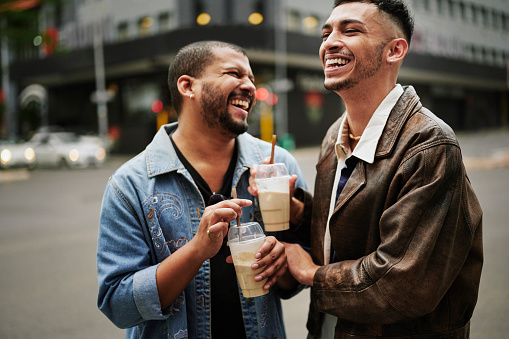 Two young male friends laughing and drinking coffee while standing together outside in the city