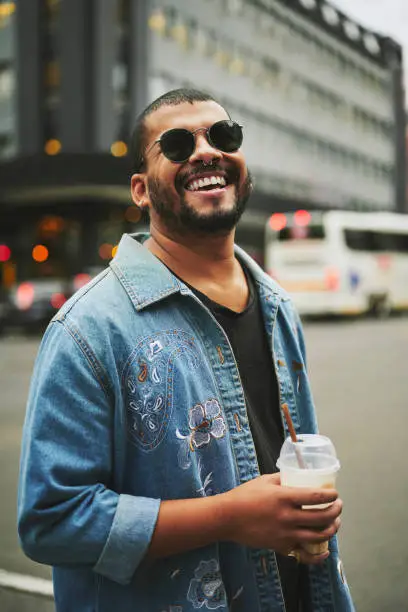 Portrait of a stylish young gay man wearing sunglasses laughing while drinking ice coffee outside on a city street