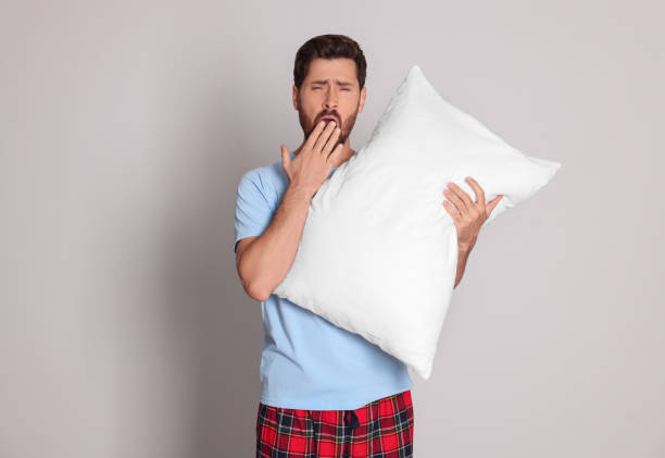 istock Tired man with pillow yawns on light grey background 1488926834