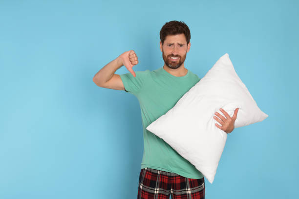 istock Sad handsome man showing thumb down and holding soft pillow on light blue background, space for text 1488926967