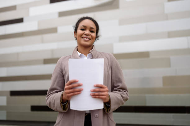 афроамериканка-менеджер в деловой одежде держит в руках документы на фоне городского пейзажа. - eadership стоковые фото и изображения