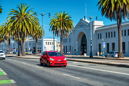 Red car drives on the palm tree lined Embarcadero street in downtown San Francisco, California, USA on a sunny day.