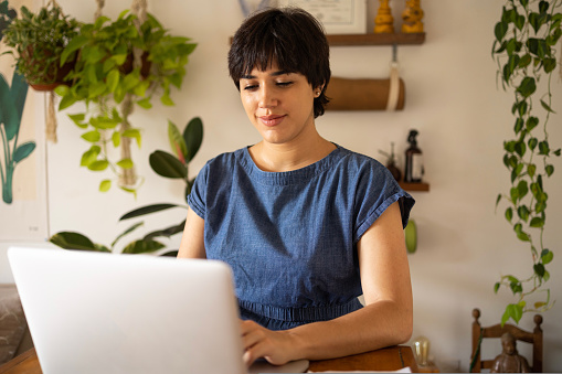 Young latin woman with short hair wearing a blue shirt. She is sitting in the apartment full of plants and using a laptop with a smile. High quality photo