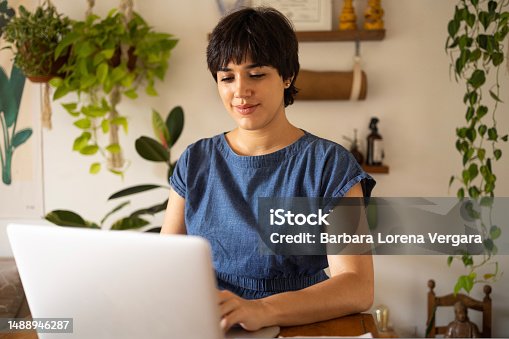 istock Young latin woman with short hair wearing a blue shirt. She is sitting in the apartment full of plants and using a laptop with a smile. 1488946287