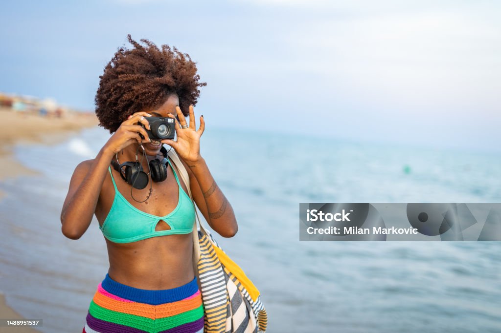 I love these moments for myself, enjoying a beautiful summer day at the sea A young black woman takes a moment for herself, enjoying a beautiful summer day at the sea, walking on a sandy beach in shallow water and capturing moments with her camera Adult Stock Photo