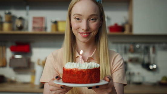 Cheerful attractive girl holding tasty cake smiling on laptop web camera closeup. Happy blond woman wearing cone bright hat blowing on candle. Young lady celebrating event with friends on video call.