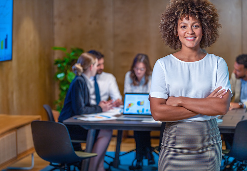 Businesswoman and team at the office. The woman is looking at camera with her business team in the background. She looks like a confident manager, business owner of entrepreneur; There are financial graphs and charts on the laptop in the background