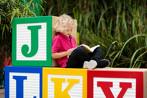 Child reading book in school yard. Kid learning abc letters. Little boy sitting on wooden toy blocks with alphabet in preschool or kindergarten. Kids read books. Children learn and study.