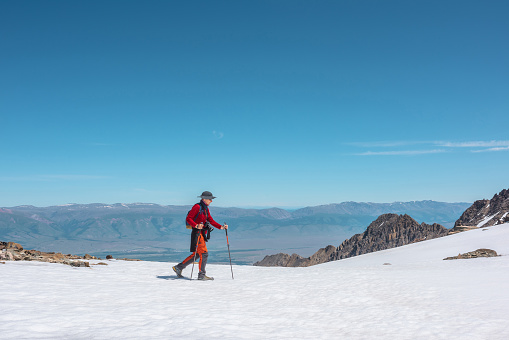 Tourist in red walks on snow mountain near abyss edge on high altitude under blue sky in sunny day. Man with camera on snowy mountain near precipice edge with view to large mountain range in away.