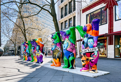 The photo captures the spirit of Berlin's Mitte District on a sunny spring day. In the center of the frame, a row of colorful bears, Berlin's symbols, stand with raised paws. Kurfürstendamm, one of the city's most bustling streets, can be seen in the background, lined with various shops and cafes.