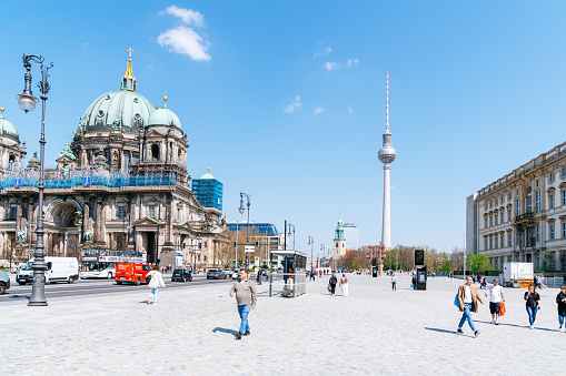 The photo captures the bustling pedestrian way along Karl-Liebknecht-Straße. The iconic Berliner Dom cathedral and the Fernsehturm tower dominate the center of the composition while St. Marienkirche can be seen towering over the city skyline in the distance.