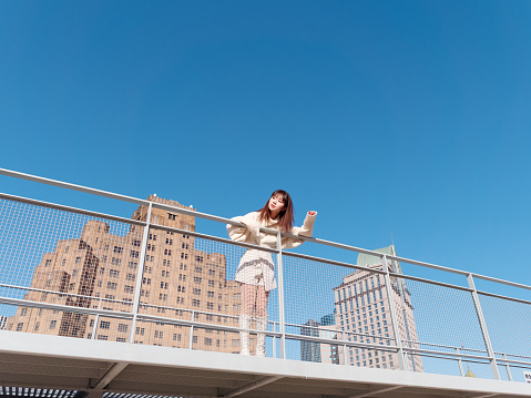 Beautiful young woman with black long hair in white skirt posing with blue sky and modern city buildings background in sunny day. Emotions, people, beauty, travel and lifestyle concept.