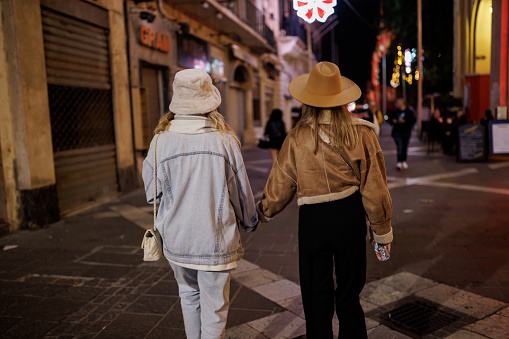 Back view of female friends holding hands and walking in city at night