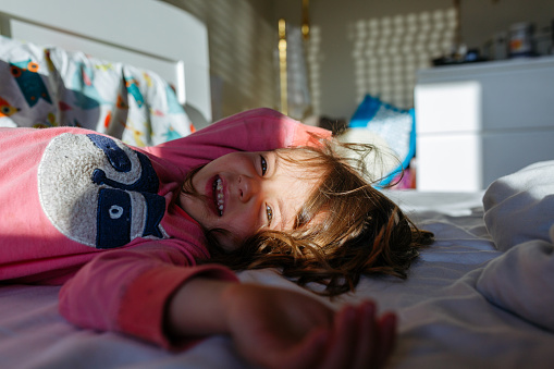 smiling little girl in pajamas lying down on parent's bed in the morning light