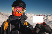 snowboarder holds an empty lift pass with a mountain in the background. Blank ski pass in the hand of a young skier in winter gear looking at the camera. Concept illustrating ski entry fee