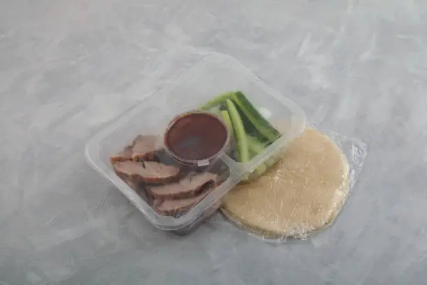 Photo of Delivery food. Peking duck in disposable plastic box. Very popular Chinese fast food. Beijing roast duck served cucumber and sweet bean sauce with pancakes