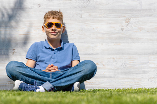 A schoolboy in sunglasses sitting on a grassy field. With copy space