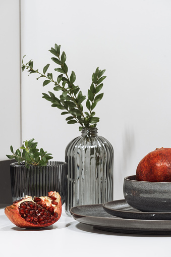 detail in decor in domestic kitchen with modern interior, composition of pomegranate, bowl, clean plates and glass vase with green twigs on table