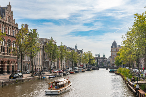 Amsterdam vintage tourboat in a canal with house boats in the canals with canal houses and green trees in the background.