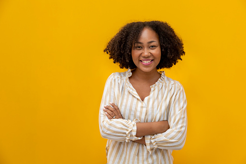 Portrait studio shot of African American woman smiling in casual cloth with big smile and crossed hand isolated on yellow background looking at camera for housewife and student confidence