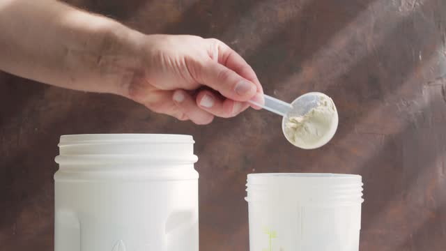 Male hand puts scoop with white whey or soy protein powder from a jar into plastic shaker, process of making protein smoothie drink, sport nutrition