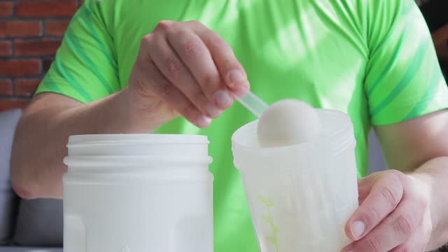 Man in sport shirt puts scoop with white whey or soy protein powder from a jar into plastic shaker, process of making protein smoothie drink, sport nutrition, cropped image