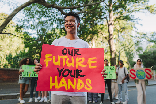 Cheerful teenage boy looking away while holding a banner sign during a climate change rally. Happy young activist protesting against global warming with a group of demonstrators in the background.