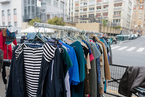 Clothes on a second-hand market in Paris, outdoors at a clothing rack, jackets, dresses and sweaters. A street and some buildings in the background.