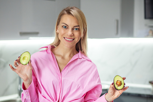 young smiling woman holding avocado in hands in kitchen. Concept of healthy food, diet and eco products.