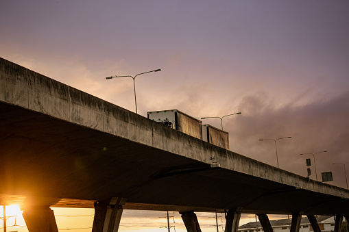 Bottom view of elevated concrete highway with truck driving. Overpass concrete road. Road flyover structure. Motorway with sunset sky. Concrete bridge engineering construction. Bridge architecture.