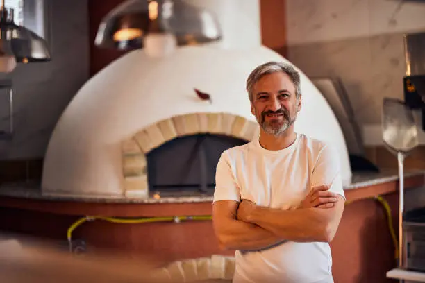 A skilled senior male pizza master with arms crossed standing in front of the pizza oven in the kitchen, smiling for the camera.