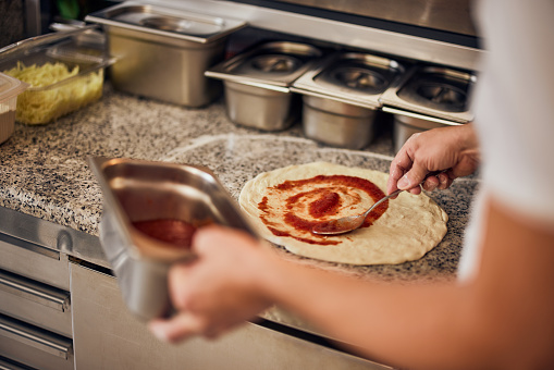 A skilled male pizza master spreads tomato sauce over the pizza dough in the restaurant's kitchen.