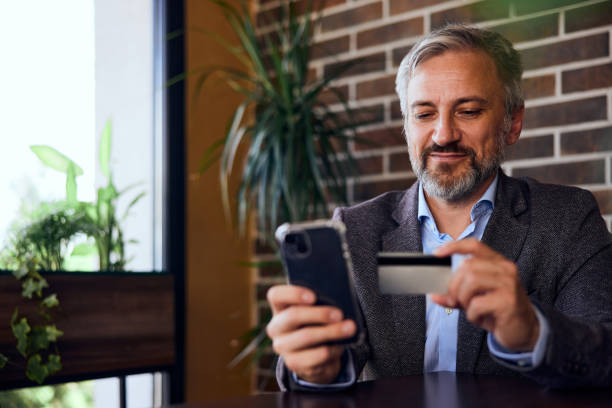 istock Smiling senior man paying over his phone, using a card while sitting at the restaurant. 1489109928