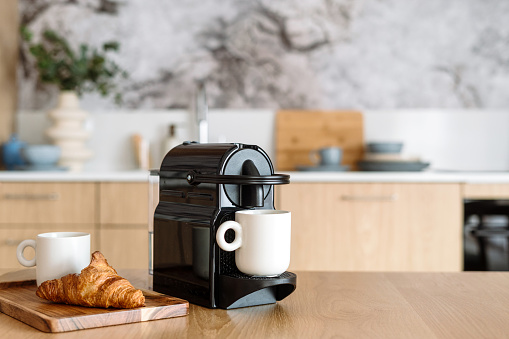 Close up view of modern espresso coffee machine with a white cup on beige table in the kitchen interior. Wooden tray with a croissant near coffeemaker.