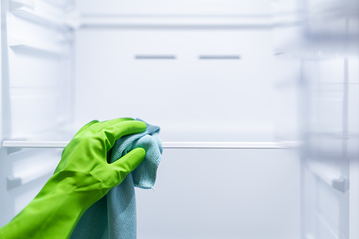 Close up view of woman hand in green rubber protective glove and a blue rag wash, clean glass refrigerator shelves. Cleaning service, housewife, routine housework.