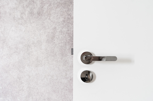 Close up view of opened modern white door with nickel handle and lock. Grey wall near the entrance. Minimalistic interior design. Copy space.