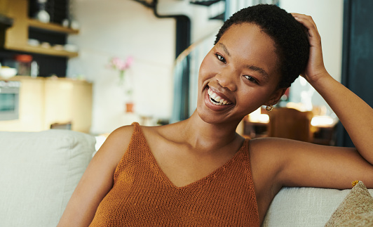 Close up shot of a beautiful young black woman sitting on her couch in her living room with her head tilted to the side smiling at the camera stock photo