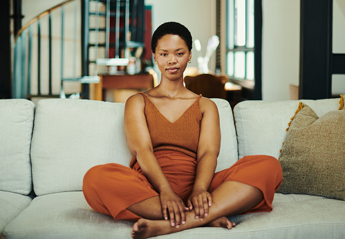 Beautiful young black woman sitting crossed legged on her couch or sofa in her lounge whilst casually dressed in a pants and top at her apartment or flat, relaxing with copy space stock photo